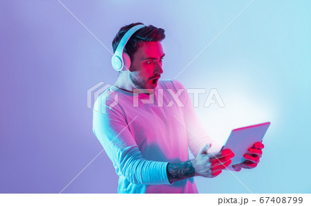 Amazing surprise online. Shocked man with open mouth in headphones holds tablet neon light 67408799