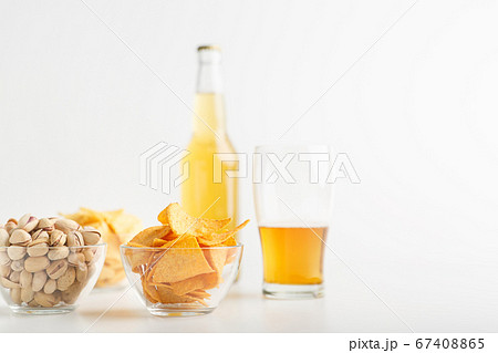 Pistachio, chips and nachos in plates. Glass of unfinished light beer and full bottle of lager 67408865
