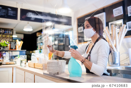 Young woman with face mask working indoors in cafe, disinfecting counter. 67410683