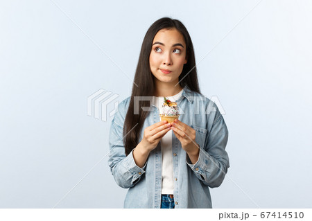 Social distancing lifestyle, covid-19 pandemic, celebrating holidays during coronavirus concept. Thoughtful cute asian girl deciding eat cupcake or note, look up thinking, holding delicious dessert Social distancing lifestyle, covid-19 pandemic, celebrating holidays during coronavirus concept. Thoughtful cute asian girl deciding eat cupcake or note, look up thinking, holding delicious dessert 67414510