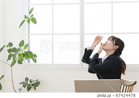 窓際の机で目薬を差すスーツ姿の若い女性 67424478