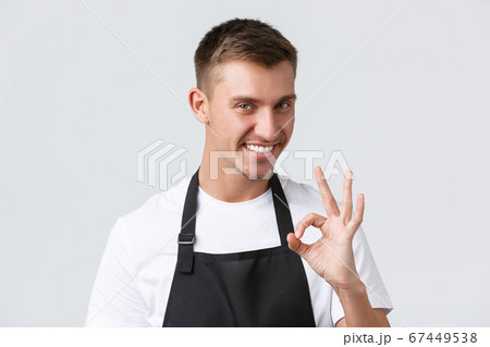 Cafe and restaurants, coffee shop owners and retail concept. Close-up of cheeky handsome waiter in black apron, barista guarantee quality of drinks, showing okay sign and smiling pleased Cafe and restaurants, coffee shop owners and retail concept. Close-up of cheeky handsome waiter in black apron, barista guarantee quality of drinks, showing okay sign and smiling pleased 67449538