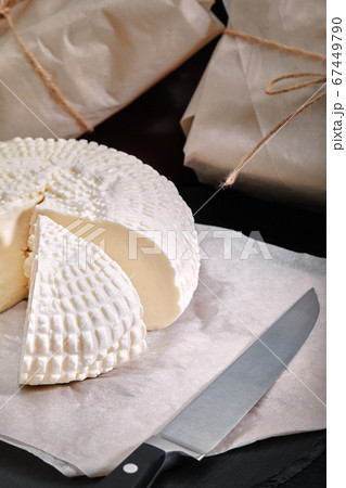 Adyghe cheese cut off a triangular piece. Nearby are three cheese heads packed in paper. On a wooden board. Background gray concrete Adyghe cheese cut off a triangular piece. Nearby are three cheese heads packed in paper. On a wooden board. Background gray concrete 67449790