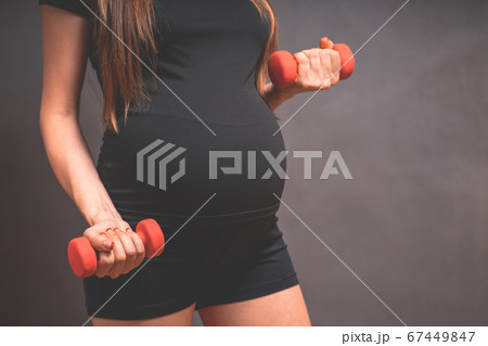 Young pregnant woman exercises with red fitball and dumbell. Working out and fitness, pregnancy Young pregnant woman exercises with red fitball and dumbell. Working out and fitness, pregnancy 67449847