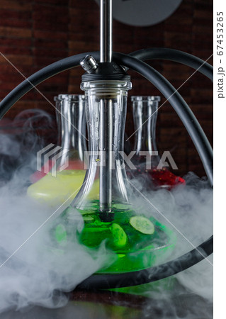Stylish oriental shisha concept. Smoking hookah on dark table, in clouds of smoke and light. Glass hookah flask with green liquid and cucumber slices. Colored flasks on smoky blurred black background. Stylish oriental shisha concept. Smoking hookah on dark table, in clouds of smoke and light. Glass hookah flask with green liquid and cucumber slices. Colored flasks on smoky blurred black background. 67453265