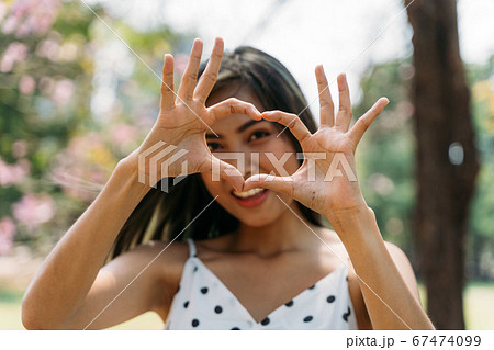 Young brunette woman with smile face making heart shape hands gesture in park. Beautiful Asian girl with toothy smile outdoors 67474099