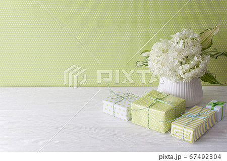 Little green gifts and a bouquet of white hydrangea in a vase. Green wall and white wooden table. Little green gifts and a bouquet of white hydrangea in a vase. Green wall and white wooden table. 67492304