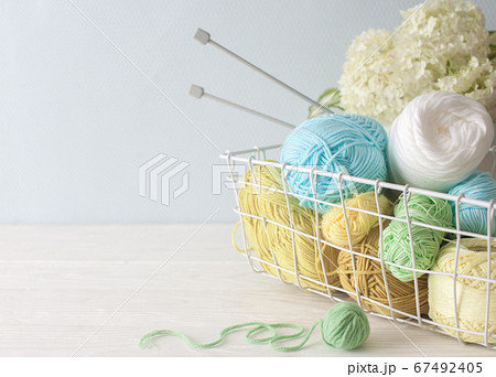 Yarn of green, yellow, blue and white colors lies in a metal basket. Hydrangea bouquet in a vase in the Nordic style. Gray scissors and knitting needles. 67492405