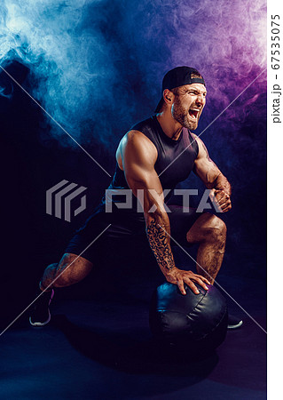 Aggressive bearded muscular sportsman is working out, push up with a medicine ball isolated on dark studio background with smoke 67535075
