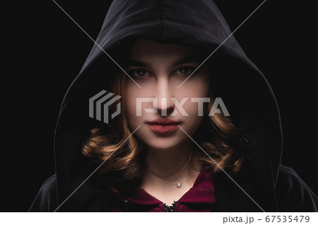 Close-up portrait of a secretive young girl in a deep dark hood on a black background. The concept of secrecy of secrets and people hiding from the government. 67535479