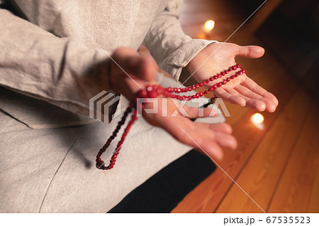 close-up man in clothes for practice and meditation sits in a lotus pose and holds red rosary to concentrate attention in a wooden room with dim light 67535523