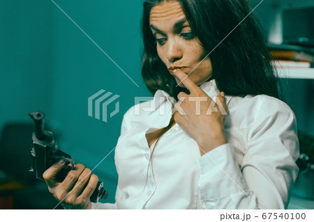 Crazy business woman with gun in office. Young beauty brunette holding gun looking on it while holding her finger on the trigger. Weapons concept. Tinted image 67540100