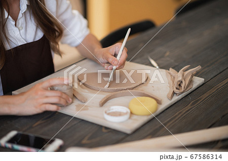 Woman's hand making clay pottery with a brush on the table with a wooden stick instrument and wooden rolling pin on it. Woman's hand making clay pottery with a brush on the table with a wooden stick instrument and wooden rolling pin on it. 67586314