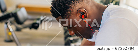 Side view of tired African American man in sportswear listening to music in wireless earphones while sitting on bench and resting during training in gym 67592794