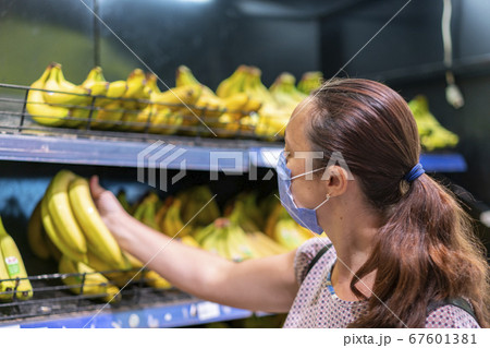 Focused woman in face mask choosing fruits, taking bananas from shelves in grocery store. Customer in supermarket. Side view. Shopping during epidemic concept 67601381