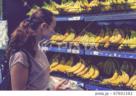 Focused woman in face mask choosing fruits, taking bananas from shelves in grocery store. Customer in supermarket. Side view. Shopping during epidemic concept. toned 67601382