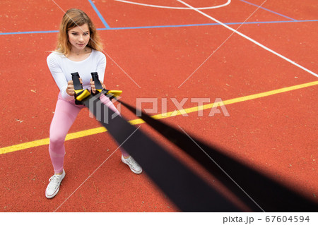 Pretty young Caucasian girl in pink sportswear exercising with suspension trainer sling, doing squats, outdoors at red sport court. Fitness concept with trx trainer Pretty young Caucasian girl in pink sportswear exercising with suspension trainer sling, doing squats, outdoors at red sport court. Fitness concept with trx trainer 67604594