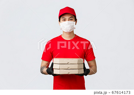 Food delivery, contactless shopping online concept. Friendly asian delivery guy in red uniform in medical mask and gloves standing clients doorstep, deliver pizzas, courier 67614173