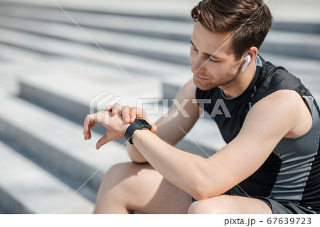 Modern jogging gadgets. Tired athlete with wireless headphones looks at fitness tracker and sits on steps 67639723