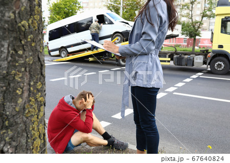 An agent fills out documents after car accident, driver sits nearby and holds his head with his hands An agent fills out documents after car accident, driver sits nearby and holds his head with his hands 67640824