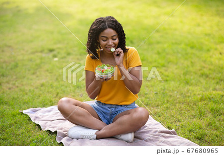 Healthy diet. Slim African American lady eating tasty vegetable salad on picnic blanket at park. Panorama 67680965