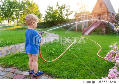 Cute adorable caucasian blond toddler boy enjoy having fun watering garden flower and lawn with hosepipe sprinkler at home backyard at sunny day. Child little helper learn gardening at summer outdoor Cute adorable caucasian blond toddler boy enjoy having fun watering garden flower and lawn with hosepipe sprinkler at home backyard at sunny day. Child little helper learn gardening at summer outdoor 67684840