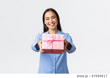 Home, holidays and lifestyle concept. Lovely smiling asian sister in blue pajama giving you gift for birthday or christmas celebration, congratulate with bday sibling, white background 67689617