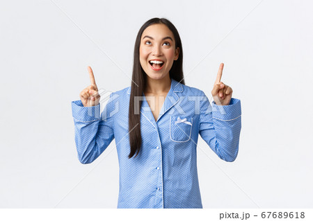 Enthusiastic attractive asian girl in blue pajamas pointing fingers up and smiling amazed as looking at awesome promo banner offer, showing announcement, react to cool new product 67689618