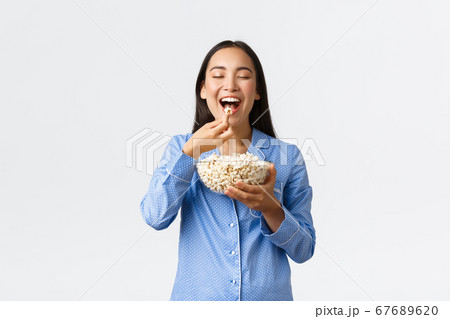 Home leisure, sleepover and slumber party concept. Smiling delighted asian girl enjoying eating favorite popcorn, wearing pajama on weekend, watching favorite movie, white background 67689620