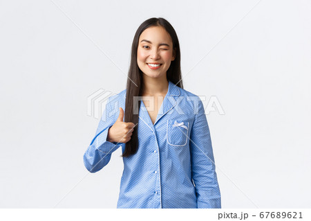 Cheerful cute asian girl in blue pajamas smiling pleased and winking at girlfriend, showing thumbs-up as praise great sleepover party, showing support, say well done or nice job, white background 67689621