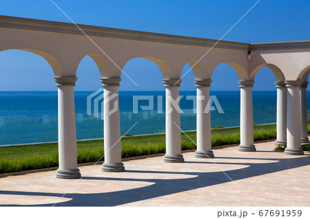 an arch with columns of white stone on sunny day and a marble floor with a lawn and a view of the sea on the coast of a luxurious mansion. 67691959