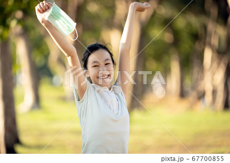 Happy smiling asian child girl is standing with raised arms up,holding mask in her hand in green nature,woman enjoy breathing fresh air after Covid-19 quarantine,Coronavirus ended,end of stay home 67700855