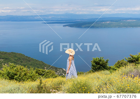 Rear view of young woman wearing striped summer dress and straw hat standing in super bloom of wildflowers, relaxing while enjoing beautiful view of Adriatic sea nature, Croatia Rear view of young woman wearing striped summer dress and straw hat standing in super bloom of wildflowers, relaxing while enjoing beautiful view of Adriatic sea nature, Croatia 67707376