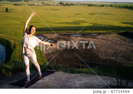 Caucasian woman practicing yoga on top of rock background of flowing river and green meadow 67714504
