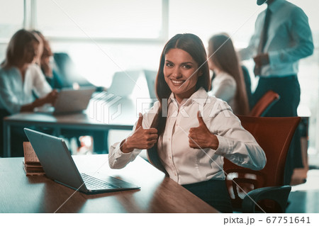 Businesswoman shows thumbs up in office. Alright gesture from young smiling girl. Group of businesspeople work on blurred background. Achievement in business concept. Tinted image Businesswoman shows thumbs up in office. Alright gesture from young smiling girl. Group of businesspeople work on blurred background. Achievement in business concept. Tinted image 67751641