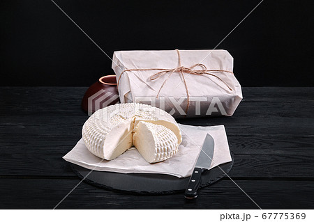 Adyghe cheese cut off a triangular piece. Nearby are three cheese heads packed in paper. On a wooden board. Background gray concrete Adyghe cheese cut off a triangular piece. Nearby are three cheese heads packed in paper. On a wooden board. Background gray concrete 67775369