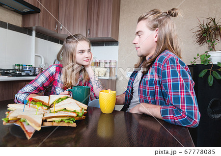 A young family drinks tea with sandwiches in the kitchen at the dinner table and looks each other. A married couple with long hair chats over tea in the home dining room 67778085