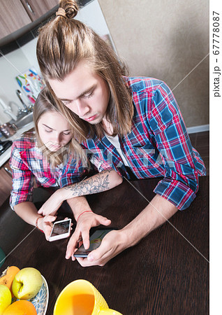 A young smile couple guy and a girl with long hair sit at the dinner table in the kitchen and use phones communicating in chat rooms and social networks. 67778087