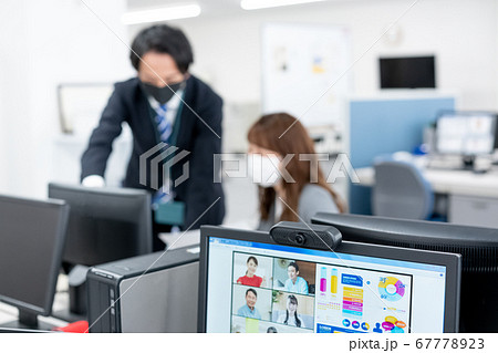 web会議用のwebカメラとその向こうにぼけるマスクをつけた会社員2人 67778923