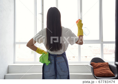 Washing windows professionally. Back view of a young woman, cleaning lady in yellow rubber gloves holding rag and spray while cleaning big window at home 67783474