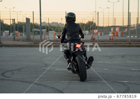 Back view of stylish woman in black leather jacket, pants and protective helmet rides on sports motorcycle at urban outdoors parking. Back view of stylish woman in black leather jacket, pants and protective helmet rides on sports motorcycle at urban outdoors parking. 67785819