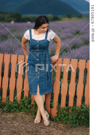 Attractive young woman walking in summer lavender field. Model dressed in denim sundress Attractive young woman walking in summer lavender field. Model dressed in denim sundress 67805191