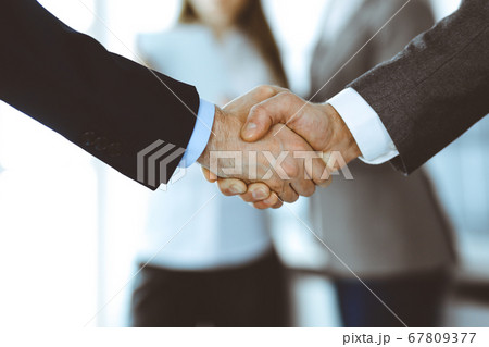 Business people shaking hands while standing with colleagues after meeting or negotiation, close-up. Group of unknown businessmen and women in modern office. Teamwork, partnership and handshake 67809377