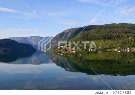 ノルウェー　ハダンゲルフィヨルド、ウルヴィク地区、早朝の朝日に輝く西側の家々、山々と水面を望む 67817882