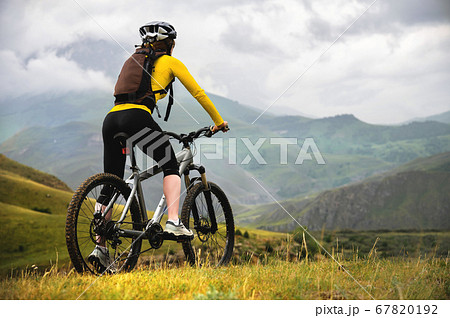 A young slender girl with a backpack and a bicycle helmet sits on a bicycle high in the mountains on a cloudy day and looks at the mountains and clouds. 67820192