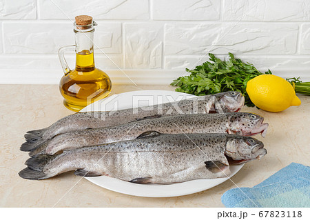 Three raw gutted trout fishes on a white plate over a kitchen table prepared for cooking. Three raw gutted trout fishes on a white plate over a kitchen table prepared for cooking. 67823118