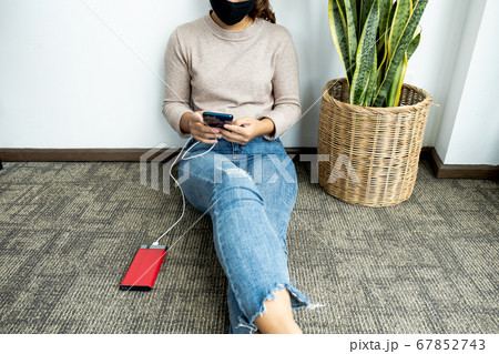 young woman sitting against a wall holding a smartphone while charging a smartphone battery young woman sitting against a wall holding a smartphone while charging a smartphone battery 67852743