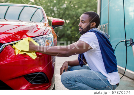 Young African man washing and wiping his red car headlight at the outdoor car wash self service. Dark skinned man with a microfiber wipe polishing the car Young African man washing and wiping his red car headlight at the outdoor car wash self service. Dark skinned man with a microfiber wipe polishing the car 67870574