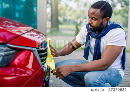 Closeup side view of red car cleaning with yellow microfiber cloths by handsome bearded African man in casual wear, wiping car radiator grille at outdoors car wash station 67870622