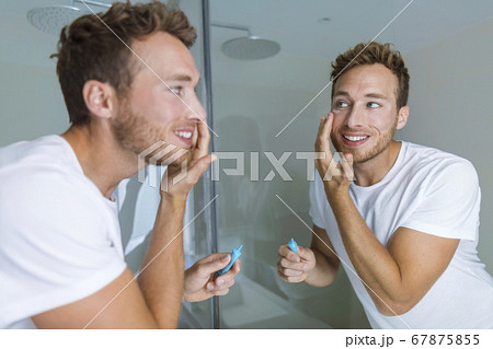 Man putting skincare facial treatment cream on face. Anti-aging skin care product. Male beauty morning routine at home lifestyle. Guy looking in bathroom mirror applying moisturizer under eyes Man putting skincare facial treatment cream on face. Anti-aging skin care product. Male beauty morning routine at home lifestyle. Guy looking in bathroom mirror applying moisturizer under eyes 67875855